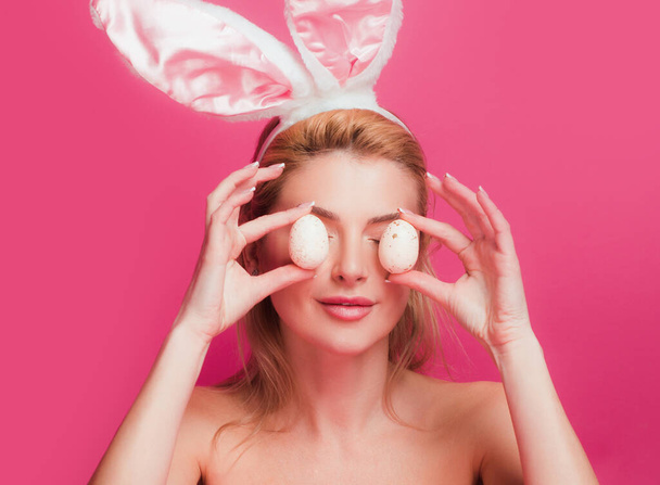 Playful young woman wearing bunny ears and holding up a colorful Easter egg in front of her eye - Fotó, kép