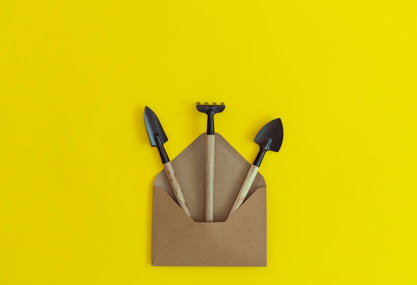 Three garden tools spill out of a craft envelope, lying on a yellow background with copy space on the sides, flat lay close-up. The concept of gardening, sowing seeds, gifts, creative. - Photo, Image
