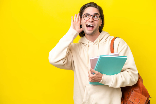 Νεαρός μαθητής όμορφος άντρας απομονωμένος σε κίτρινο φόντο ακούγοντας κάτι βάζοντας το χέρι στο αυτί. - Φωτογραφία, εικόνα