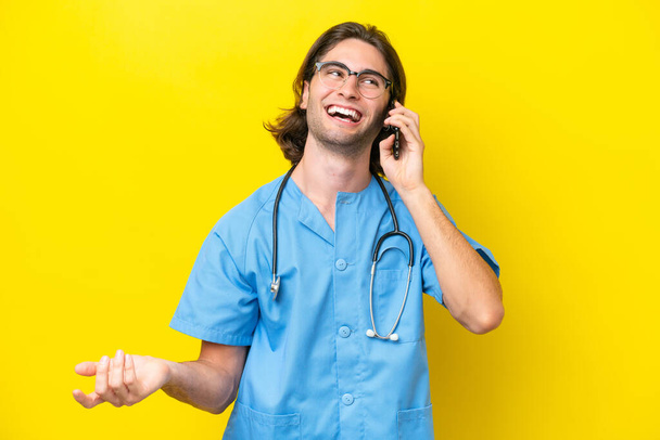 Fiatal sebész kaukázusi férfi elszigetelt sárga alapon tartja a beszélgetést a mobiltelefon valakivel - Fotó, kép