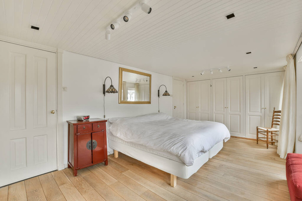 a bedroom with wood flooring and white walls, there is a red chair in the room next to the bed - Photo, image