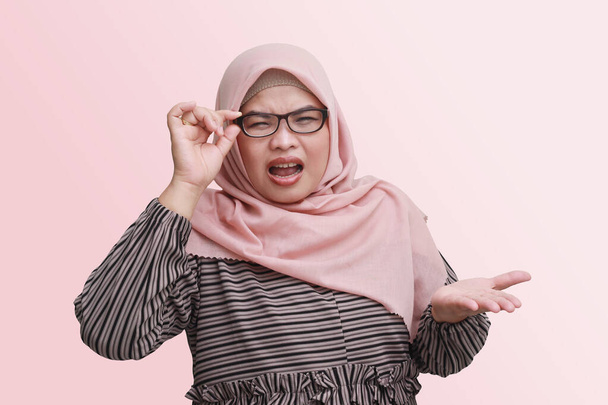 Portrait of unpleasant Asian woman with hijab, adjusting eyeglasses, checking her sight problem and showing confusion gesture with hands. Isolated image on pink background - Photo, Image