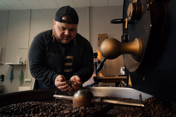 Άνδρας εργάτης με καπέλο και μαύρη στολή στέκεται κοντά στη μηχανή ψησίματος με μια χούφτα κόκκους καφέ και μυρίζει καβουρδισμένους κόκκους - Φωτογραφία, εικόνα