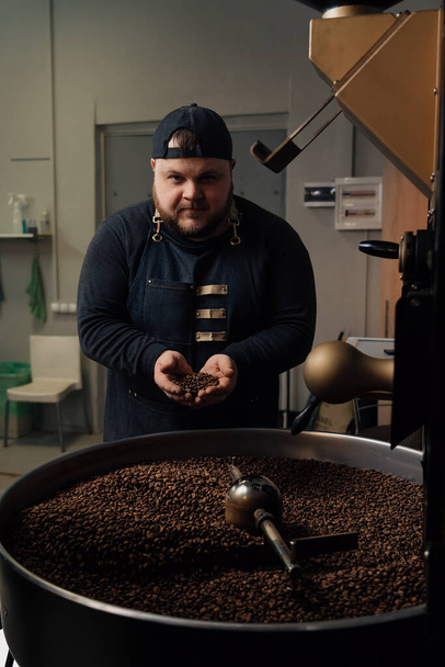 Maschio lavoratore in berretto e uniforme nera in piedi vicino torrefazione macchina con una manciata di chicchi di caffè e grani tostati odore - Foto, immagini