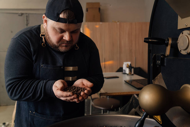 Travailleur masculin en casquette et uniforme noir debout près de la machine de torréfaction avec une poignée de grains de café et l'odeur des grains torréfiés - Photo, image