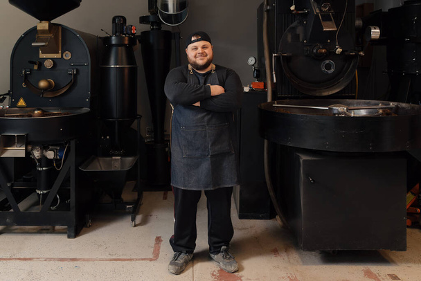 Full body of confident male worker in uniform and cap standing with crossed arms near coffee roasting machine while working at factory - Photo, Image