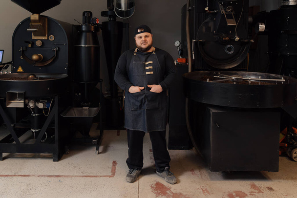 Πλήρες σώμα των ανδρών αυτοπεποίθηση εργαζόμενος σε ομοιόμορφη και καπάκι στέκεται με σταυρωμένα χέρια κοντά σε μηχανή καφέ ψήσιμο, ενώ εργάζονται στο εργοστάσιο - Φωτογραφία, εικόνα