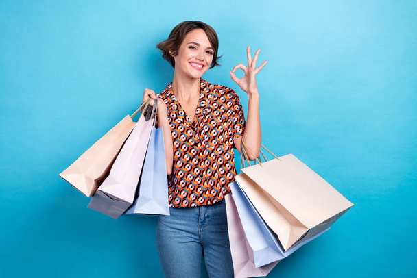 Photo of pretty shiny woman wear print blouse holding bargains showing okey gesture isolated blue color background. - Fotoğraf, Görsel