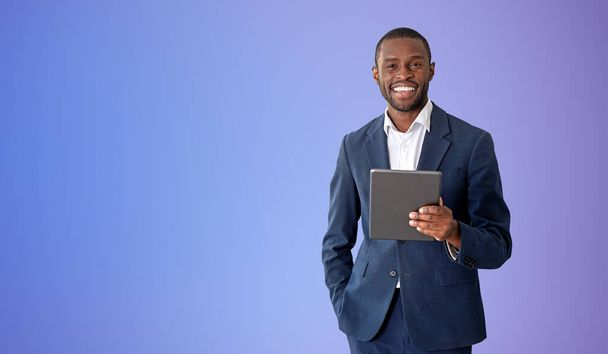 Cheerful young African American businessman holding tablet computer over purple background. Concept of social media. Mock up - Fotografie, Obrázek