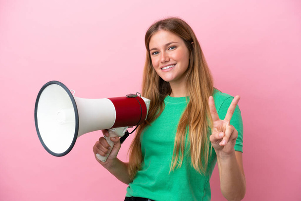 Young blonde woman isolated on pink background holding a megaphone and smiling and showing victory sign - Foto, Imagem