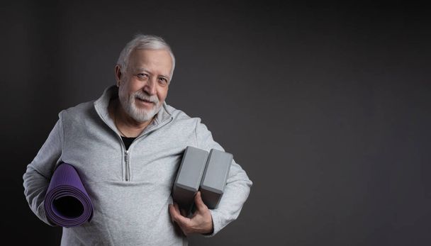 Charismatic elderly gray-haired man in tracksuit, mat and yoga support blocks, smiling in studio, gray background, space for text - Фото, зображення