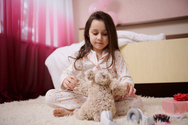 Gelukkig klein meisje, gekleed in stijlvolle pyjama, spelend met een pluche speeltje, zittend op het tapijt in haar kinderlijke kamer na dagdroom. Internationale Kinderdag en zorgeloos kinderconcept - Foto, afbeelding