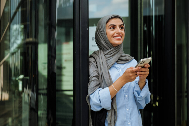Cheerful middle-aged muslim woman wearing hijab using phone while standing outdoors - Valokuva, kuva