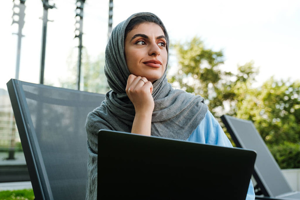 Young muslim woman wearing headscarf using laptop while sitting on chair outdoors - Foto, imagen