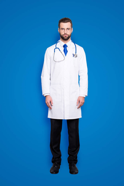 Full size portrait of attractive, concentrated virile harsh doc with beard in white lab coat, tie and stethoscope on his neck, looking at camera, isolated on grey background. - Foto, afbeelding