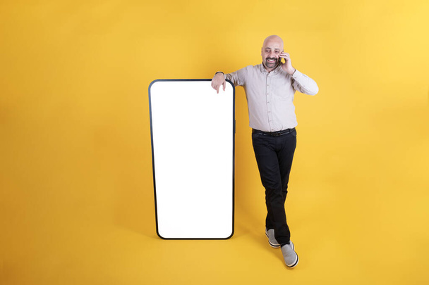 Middle aged caucasian man leaning big smartphone with empty white blank screen for mock up. Speaking on the phone. Casual dressed small-bellied guy standing over yellow background, copy space. - Photo, Image