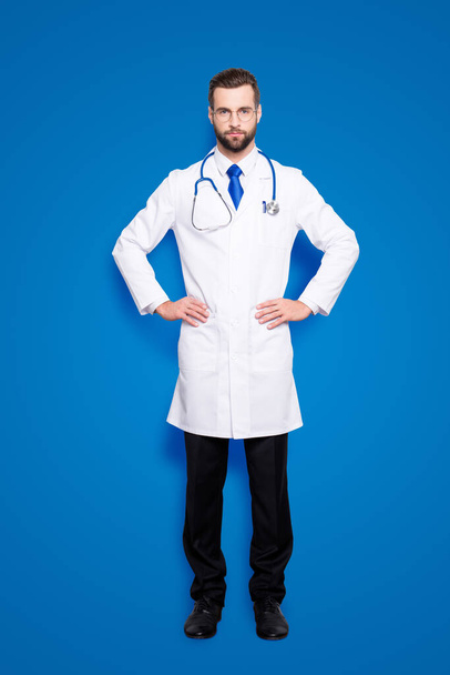 Full size portrait of attractive concentrated doc with stubble in white lab coat, tie and stethoscope on his neck, holding arms on waist, looking at camera, isolated on grey background. - Valokuva, kuva