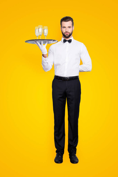 Full size fullbody portrait of concentrated man in classic white shirt and black bowtie holding hand behind the back and tray with three glasses of sparkling wine, isolated on grey background. - Photo, image