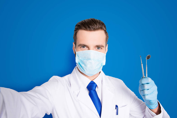 Self portrait of successful cheerful dentist in white lab coat, blue tie, shooting selfie on front camera, holding tools, having leisure, fun, video call, isolated on grey background. - Фото, зображення