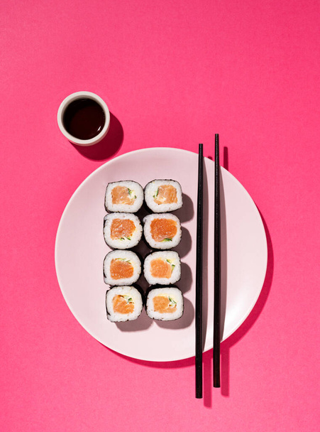 Rollo de sushi japonés con salmón, pepino y salsa de soja con palillos en el plato sobre fondo rosa con sombra dura. Concepto de fotografía de alimentos. Vista superior y espacio de copia. - Foto, Imagen