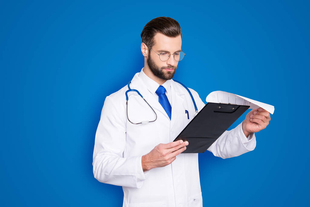 Portrait of concentrated busy doc in white lab coat and stethoscope on his neck examine, holding clipboard checking, expertising, analysing disease history, isolated on grey background. - Fotó, kép