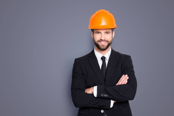Portrait of attractive young stylish investor with stubble in orange hard hat and black tux, tuxedo with tie, looking at camera, isolated on grey background, having his arms crossed. - Valokuva, kuva