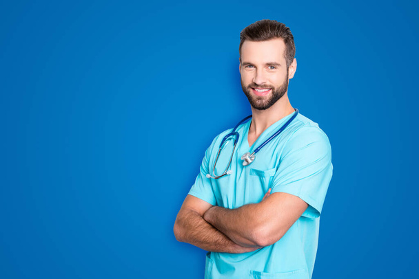 Portrait with copy space, empty place of positive cheerful man with stethoscope on his neck in blue lab uniform, having his arms crossed, isolated on grey background. - Zdjęcie, obraz