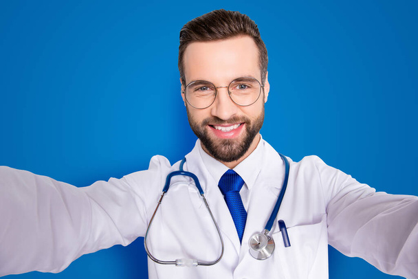 Self portrait of joyful cheerful doc in white outfit with tie and stubble having stethoscope on his neck shooting selfie with two arms, isolated over grey background. - Zdjęcie, obraz