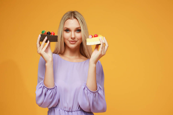 Beautiful blonde girl in a lilac dress with cakes on a yellow background. Studio. Diet and health - Photo, Image