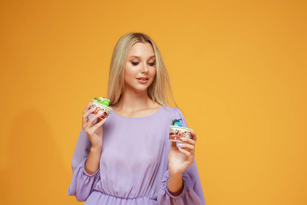 Mooi blond meisje in een lila jurk met taarten op een gele achtergrond. Een studio. Dieet en gezondheid - Foto, afbeelding