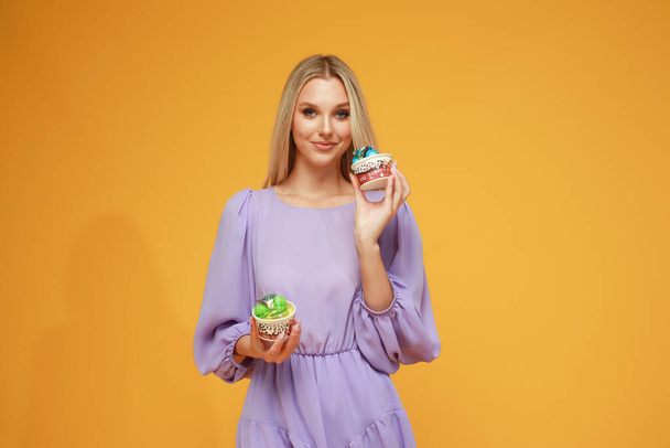 Beautiful blonde girl in a lilac dress with cakes on a yellow background. Studio. Diet and health - Фото, изображение