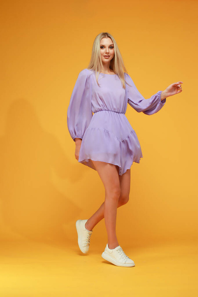 Beautiful blonde girl in a lilac dress on a yellow background. Studio. Smile and emotions. - Fotó, kép