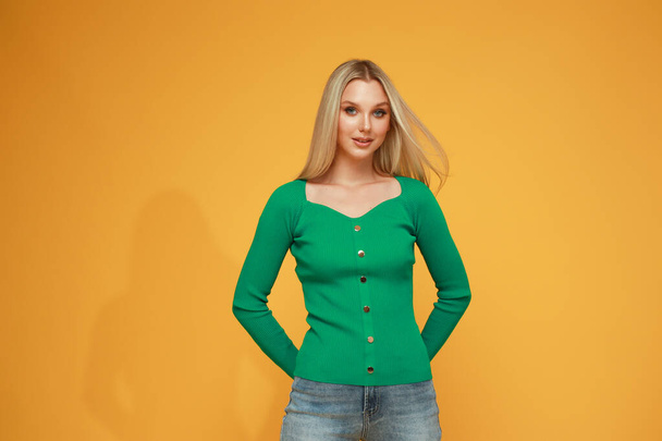 Beautiful blonde girl in a green sweater and jeans on a yellow background. Studio. Smile and emotions. - Foto, Imagem