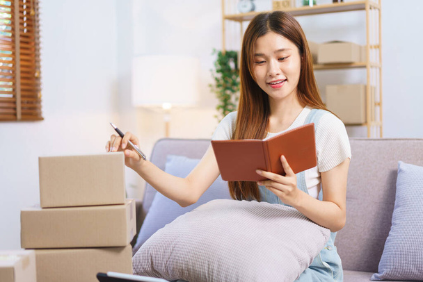 Online seller concept, Female entrepreneur writes on parcel box after reading address on notebook. - Photo, image