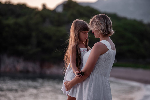Close-up portrait of pregnant mother with little daughter walking hand in hand in white dresses at sunset by the sea. Child care. Healthy motherhood. The concept of vacation, travel single parent. - Fotoğraf, Görsel