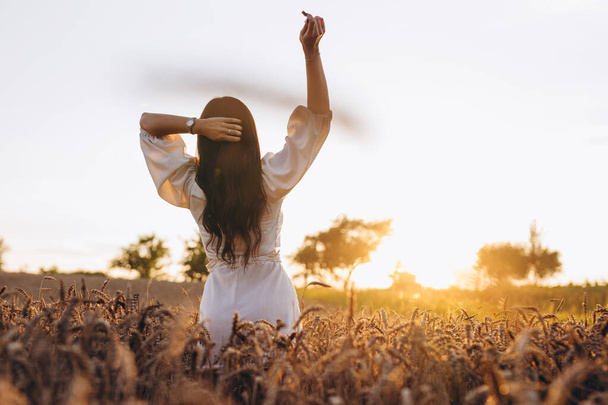 A gyönyörű hölgy a kezével pózol és nézi a naplementét. Fehér inget visel és fantasztikusan néz ki. Látjuk a napsugarakat. Van egy nagy kukoricamező a lány mellett.. - Fotó, kép