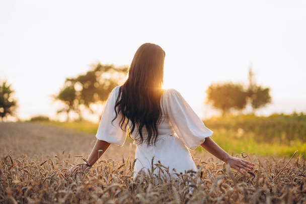 A girl is standing in the field and holding her hands out while looking at sun. She is wearing a white shirt and is looking amazing. We can see rays of sun. There is a big cornfield behind the girl. - Foto, Imagem
