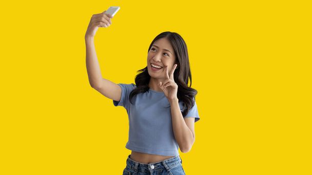 Joyeux jeune femme prenant un selfie avec un téléphone portable et levant deux doigts, Portrait de fille heureuse avec le visage lumineux isolé sur fond jaune, Prendre une photo. - Photo, image