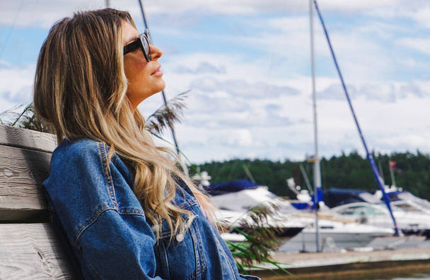Vrouw met een zonnebril die wegkijkt terwijl ze op de bank in de haven zit  - Foto, afbeelding