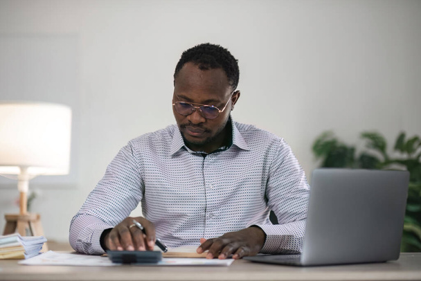 Skupiony afrykański biznesmen, trzymający dokumenty przeglądający laptopa robiący badania. koncepcja biznesowego analityka finansowego. - Zdjęcie, obraz
