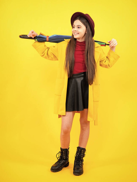 Child girl with rainbow umbrella in autumn weather isolated on yellow background. Autumn kids clothes. Smiling girl in autumn coat. Happy teenager portrait full length - Foto, imagen