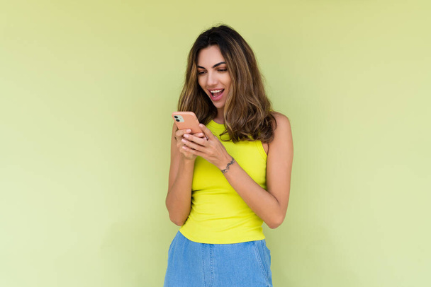 fiatal nő alkalmi kopás elszigetelt zöld háttér nézi a telefon képernyőjén nagy mosoly olvasás üzenet - Fotó, kép