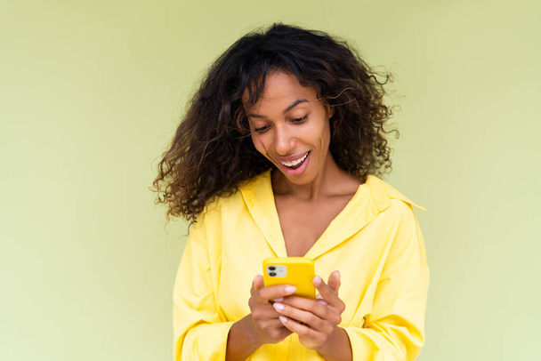 Piękna Afroamerykanka w luźnej koszuli na zielonym tle trzyma telefon komórkowy z uśmiechem - Zdjęcie, obraz