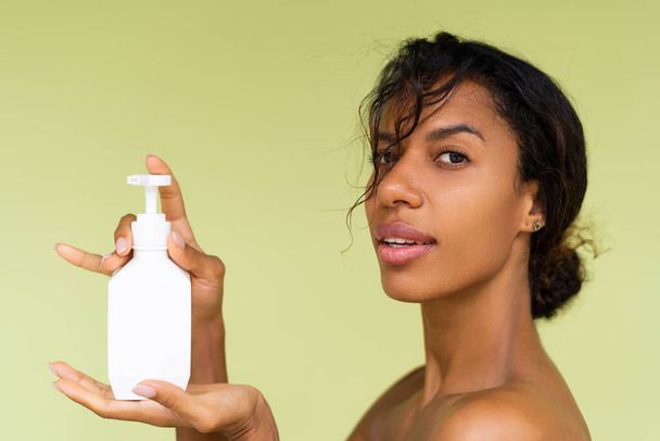 Retrato de belleza de una joven afroamericana en topless con hombros desnudos sobre fondo verde con una botella blanca de loción corporal - Foto, imagen