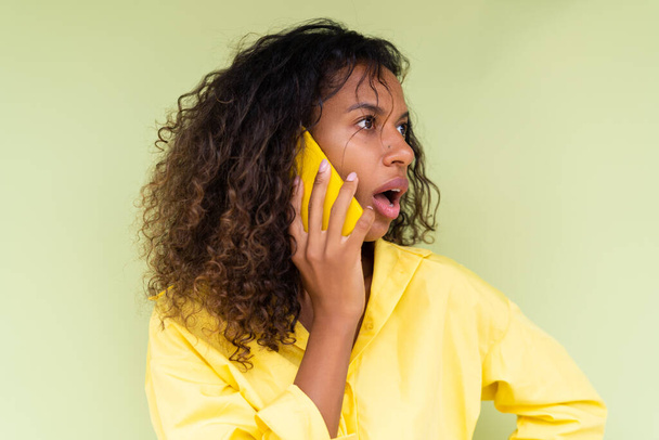 Schöne afrikanisch-amerikanische Frau in lässigem Hemd auf grünem Hintergrund Gespräch auf Handy schockiert verwirrt - Foto, Bild