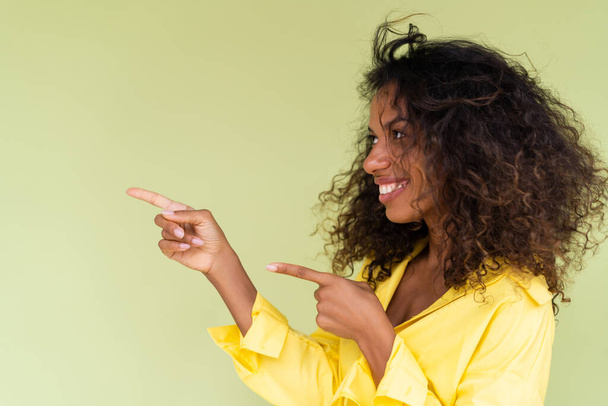 Beautiful african american woman in casual shirt on green background happy positive excited point finger to the left - 写真・画像