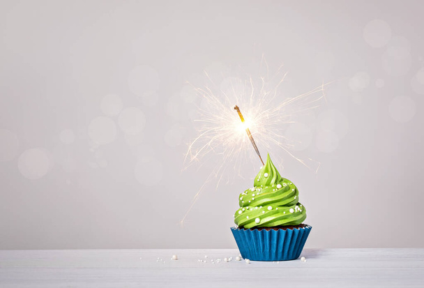 Cupcake de aniversário verde com cobertura de creme de manteiga, forro de bolo de xícara azul, polvilhas e sparkler iluminado em um fundo branco cinza claro. espaço de cópia. - Foto, Imagem