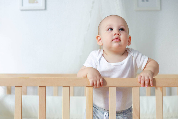 Portrait of a cute baby boy standing in his crib. Smiling happy little toddler boy stand in the baby cot - Zdjęcie, obraz