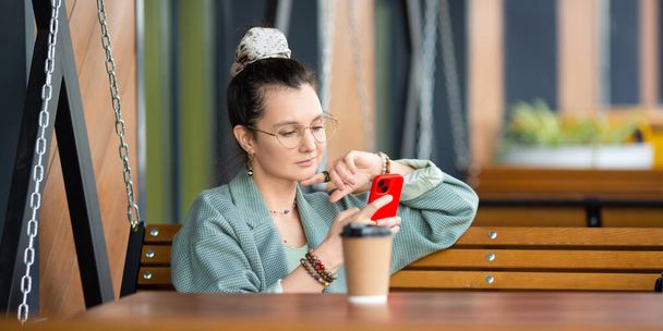 Mladá žena v brýlích si prohlíží smartphone a popíjí kávu ve venkovní kavárně. Tato stock fotografie zachycuje moderní městský životní styl se spoléháním na technologii a pohodlí. - Fotografie, Obrázek