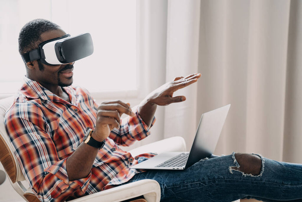 Smiling african american man in VR glasses sits in armchair with laptop, testing virtual reality goggles. Modern black guy viewing purchase in cyberspace. Tech and e-commerce concept. - Photo, image